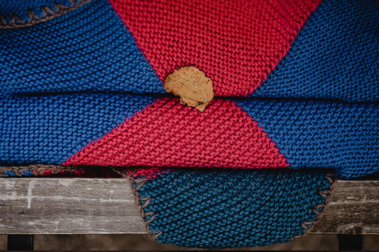 Services Handmade knitted blanket in vibrant red and blue featuring a fallen leaf on a wooden bench.